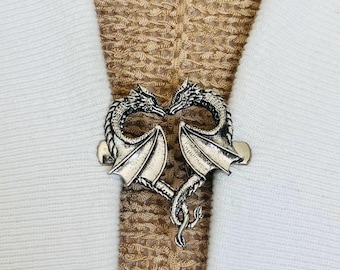 Peut inclure: Un fermoir de pull en métal argenté représentant deux dragons se faisant face, formant un cœur. Les dragons ont des ailes et des écailles détaillées. Le fermoir est fixé à un tissu blanc et beige.