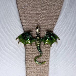 May include: A gold dragon brooch with green enamel wings and body, detailed with small crystals. The dragon is positioned on a textured beige fabric and white garment.