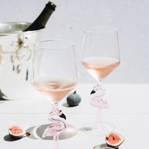 May include: Two wine glasses filled with ros&eacute;, featuring pink flamingo-shaped stems. The glasses sit on a white surface, accompanied by figs, a bottle of champagne in a silver bucket, and a cork.