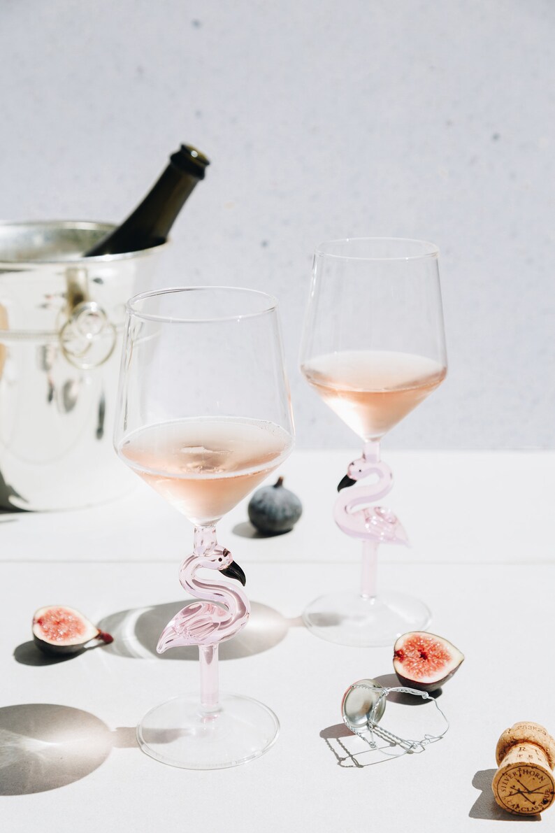 May include: Two wine glasses filled with ros&eacute;, featuring pink flamingo-shaped stems. The glasses sit on a white surface, accompanied by figs, a bottle of champagne in a silver bucket, and a cork.