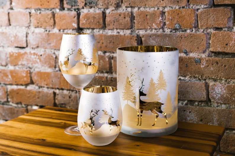 May include: Three frosted glass candle holders with gold accents and a winter scene design featuring deer and trees. The candle holders are on a wooden surface.