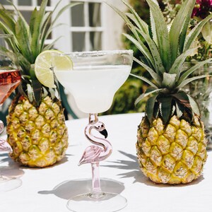 May include: Two pink flamingo margarita glasses with a lime wedge garnish, sitting on a white table with two pineapples in the background.