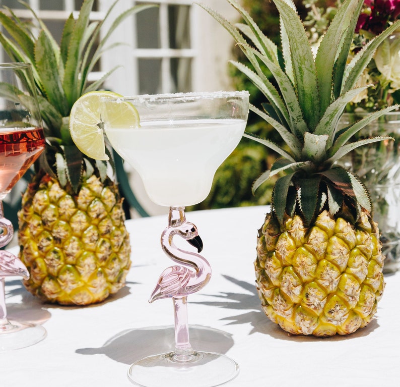May include: Two pink flamingo margarita glasses with a lime wedge garnish, sitting on a white table with two pineapples in the background.