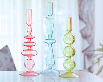 Puede incluir: Tres candelabros de vidrio en diferentes colores y estilos. Los candelabros están hechos de vidrio transparente con una variedad de formas y tamaños. Los candelabros están dispuestos en una fila sobre una superficie blanca.