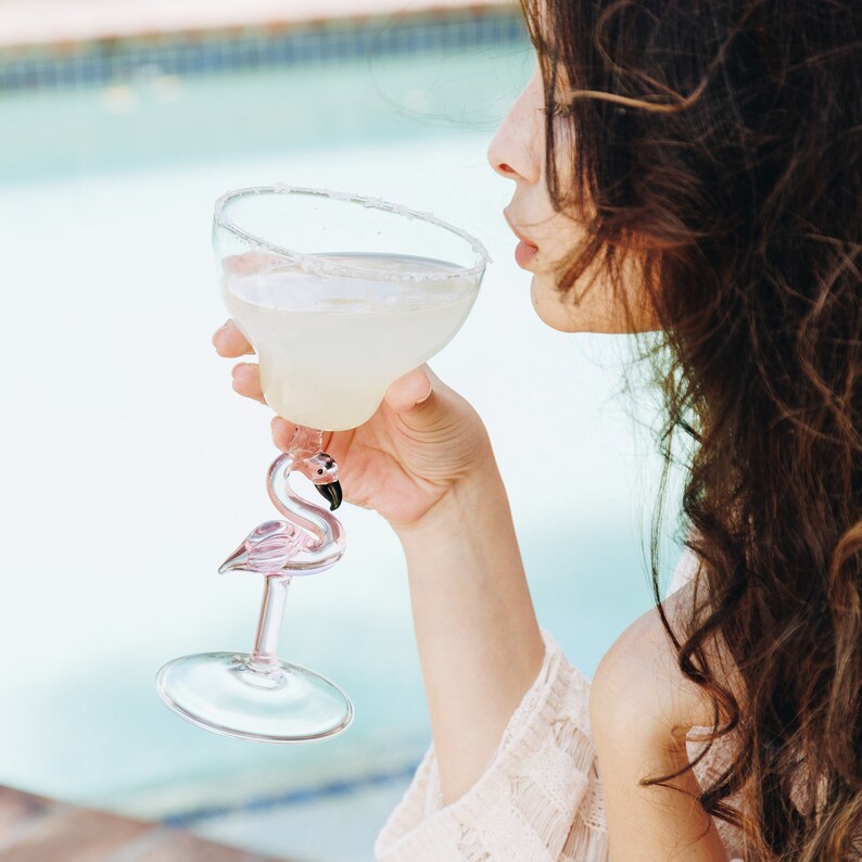 May include: A woman is holding a pink flamingo-shaped margarita glass filled with a light yellow beverage. The glass has a salted rim.