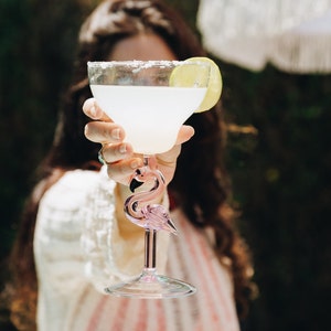 May include: A clear glass margarita glass with a pink flamingo stem, filled with a light yellow margarita and a lime wedge garnish.