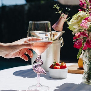 May include: A clear wine glass filled with pink wine, featuring a pink flamingo-shaped stem. The glass is held by a hand, with a background of strawberries, flowers, and a champagne bottle in an ice bucket. The scene is set outdoors.