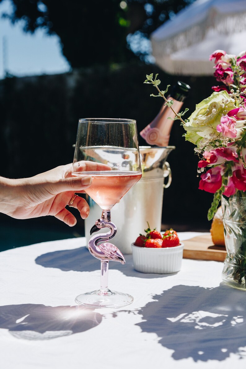 May include: A clear wine glass filled with pink wine, featuring a pink flamingo-shaped stem. The glass is held by a hand, with a background of strawberries, flowers, and a champagne bottle in an ice bucket. The scene is set outdoors.
