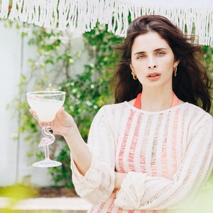 May include: A woman wearing a white and pink striped sweater holds a margarita in a flamingo-shaped glass.