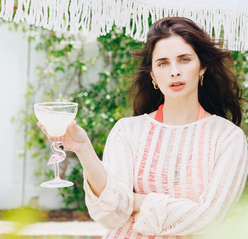 May include: A woman wearing a white and pink striped sweater holds a margarita in a flamingo-shaped glass.