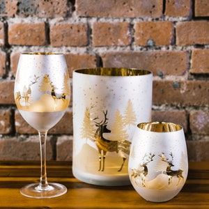 May include: Three glass candle holders with a white frosted finish and gold accents. Each holder features a winter scene with a deer and trees. The holders are arranged on a wooden surface.