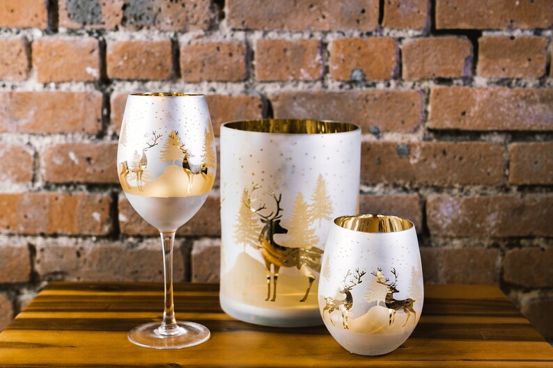 May include: Three glass candle holders with a white frosted finish and gold accents. Each holder features a winter scene with a deer and trees. The holders are arranged on a wooden surface.