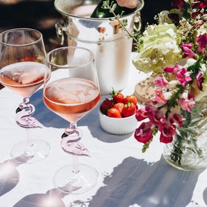 May include: Two wine glasses filled with pink wine, featuring flamingo-shaped stems, sit on a white tablecloth. A silver ice bucket, a vase of flowers, and bowls of strawberries and raspberries complete the outdoor setting.