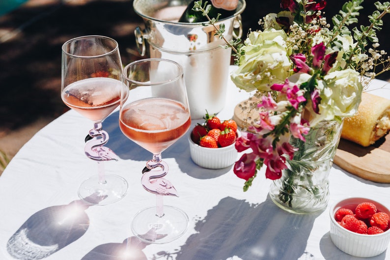 May include: Two wine glasses filled with pink wine, featuring flamingo-shaped stems, sit on a white tablecloth. A silver ice bucket, a vase of flowers, and bowls of strawberries and raspberries complete the outdoor setting.
