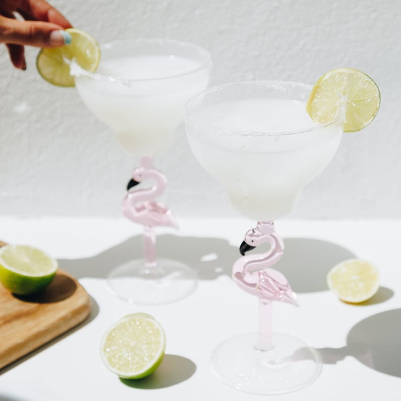 May include: Two pink flamingo margarita glasses filled with a white, frothy drink. Each glass has a lime wedge garnish.