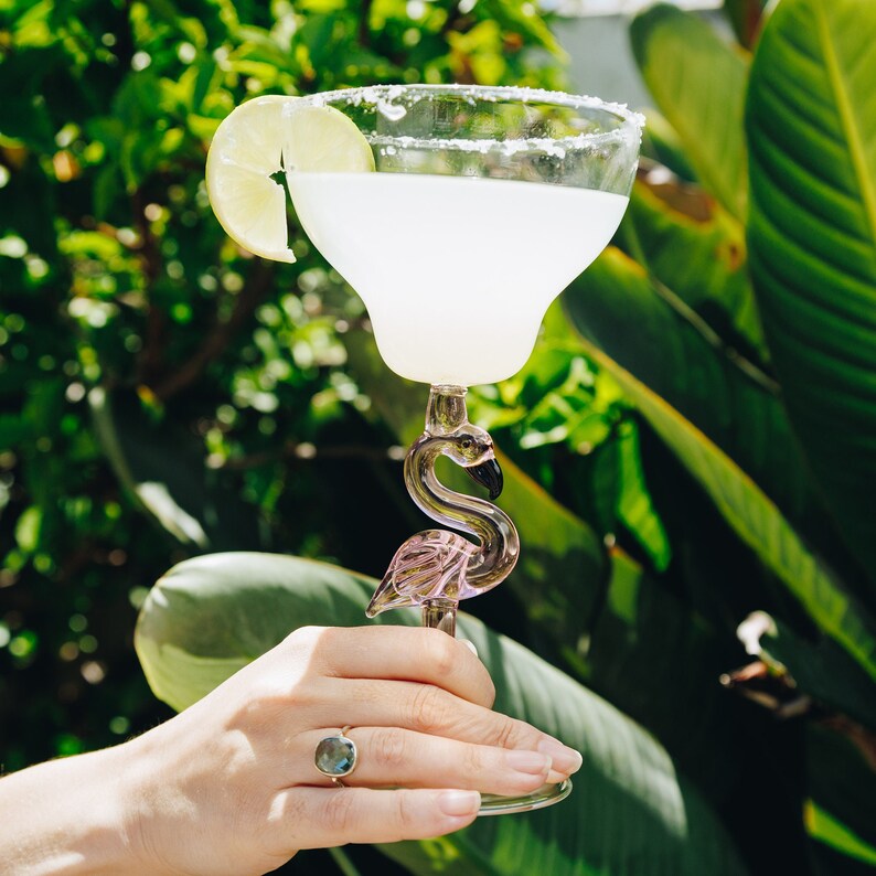 May include: A pink flamingo-shaped glass filled with a white margarita cocktail garnished with a lime wedge. The glass is sitting on a hand with a ring on the finger.