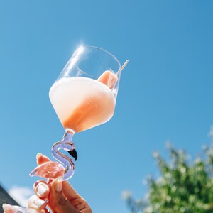 May include: A clear glass cocktail glass with a flamingo-shaped stem, filled with a pink drink and a fruit garnish. The glass is held up against a bright blue sky, with a few green trees in the background.