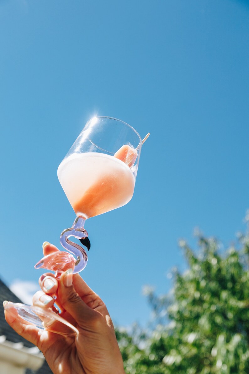 May include: A clear glass cocktail glass with a flamingo-shaped stem, filled with a pink drink and a fruit garnish. The glass is held up against a bright blue sky, with a few green trees in the background.