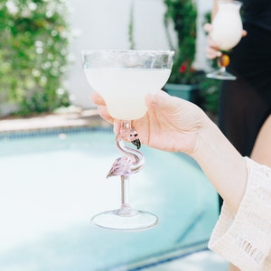 May include: A pink flamingo-shaped glass filled with a clear, light yellow beverage. The glass is being held by a person's hand, with a swimming pool in the background.