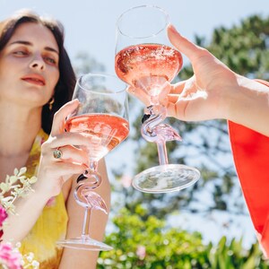 May include: Two clear wine glasses filled with pink liquid are raised in a toast. The glasses have a flamingo design on the stem. The background is a sunny outdoor scene with greenery and a blue sky.