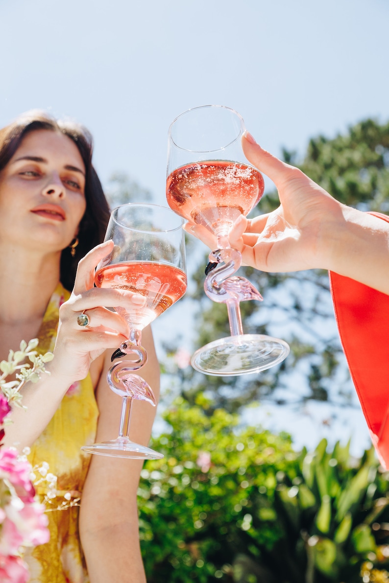 May include: Two clear wine glasses filled with pink liquid are raised in a toast. The glasses have a flamingo design on the stem. The background is a sunny outdoor scene with greenery and a blue sky.