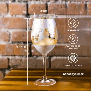 May include: A frosted glass wine glass with a gold rim and a winter scene design featuring a snowy forest and deer. The glass is 9.5 inches tall and 3 inches in diameter. It has a 19 ounce capacity and is lead-free and waterproof.