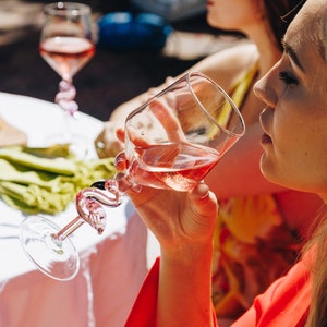 May include: A clear wine glass with a pink flamingo-shaped stem, filled with pink wine. The glass is held by a person wearing a red top. Another person is in the background, wearing a white hat and yellow top. The scene is outdoors.