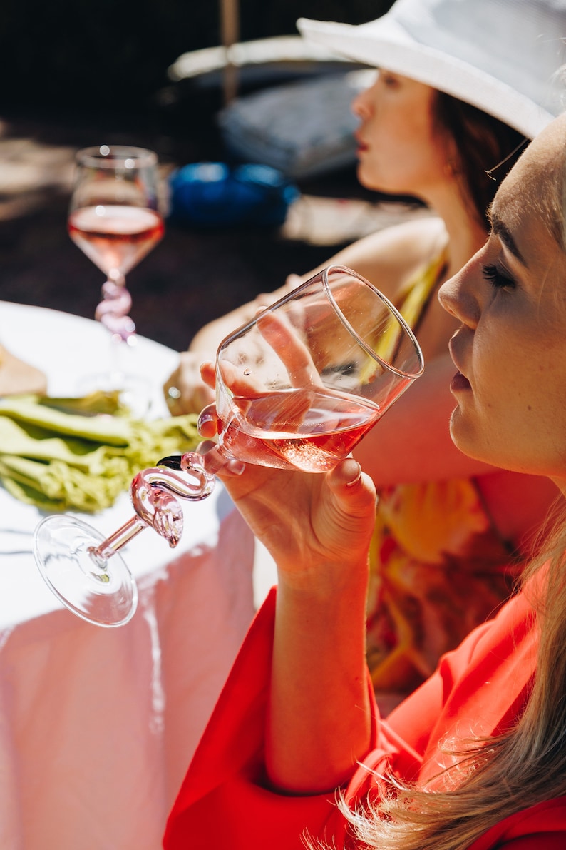 May include: A clear wine glass with a pink flamingo-shaped stem, filled with pink wine. The glass is held by a person wearing a red top. Another person is in the background, wearing a white hat and yellow top. The scene is outdoors.