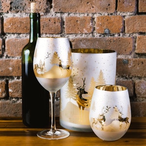 May include: Three frosted glass candle holders with a gold rim and a winter scene design featuring trees and reindeer. The candle holders are on a wooden surface.