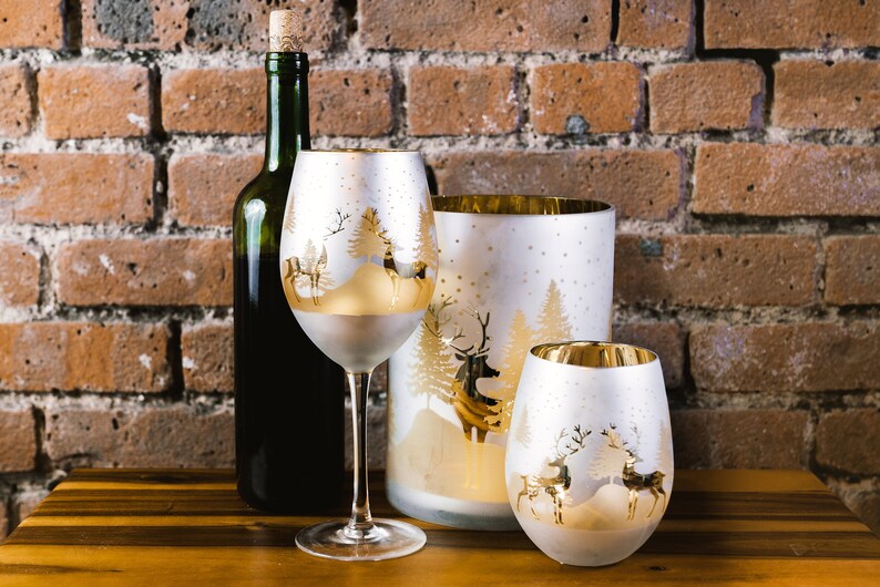 May include: Three frosted glass candle holders with a gold rim and a winter scene design featuring trees and reindeer. The candle holders are on a wooden surface.