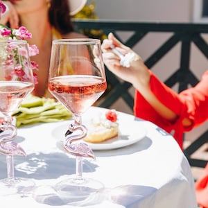 May include: Two clear wine glasses filled with pink liquid, featuring flamingo-shaped stems. The glasses sit on a white tablecloth, with flowers and a dessert plate in the background. The glasses are part of a set.