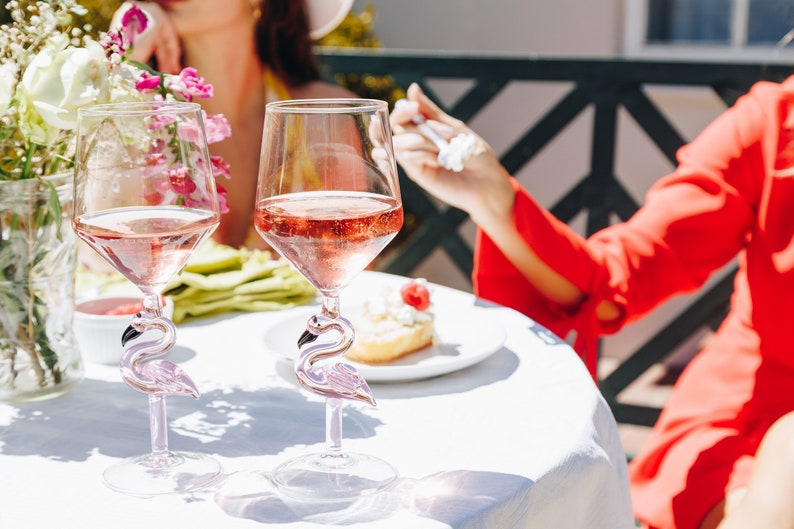 May include: Two clear wine glasses filled with pink liquid, featuring flamingo-shaped stems. The glasses sit on a white tablecloth, with flowers and a dessert plate in the background. The glasses are part of a set.