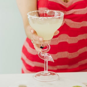 May include: A clear glass margarita glass with a salted rim, filled with a light yellow margarita, sits on a flamingo-shaped glass stem. The glass is being held by a person wearing a red and white striped dress.