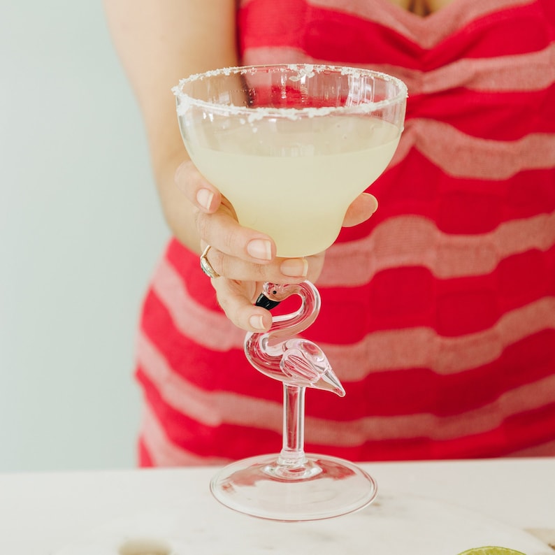 May include: A clear glass margarita glass with a salted rim, filled with a light yellow margarita, sits on a flamingo-shaped glass stem. The glass is being held by a person wearing a red and white striped dress.