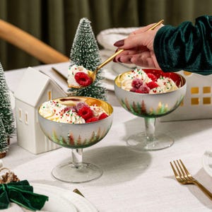 May include: Two dessert bowls filled with whipped cream, raspberries, and sprinkles. The bowls have a silver exterior with a gold interior and a forest design. A gold spoon is scooping a portion of the dessert. Christmas-themed.