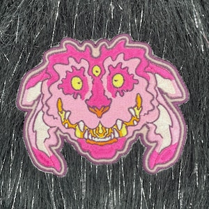May include: Pink and yellow embroidered patch of a creature with a toothy grin and three eyes. The patch is on a black fuzzy background.