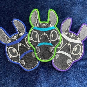 May include: Three embroidered patches of animal faces with black and white stripes and different colored outlines. The patches are green, blue, and purple.