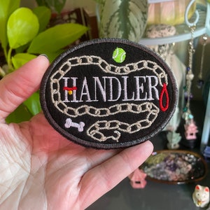 K&ouml;nnte beinhalten: Ein schwarzer gestickter Patch mit einem wei&szlig;en Kettenmuster und dem Wort "HANDLER" in wei&szlig;er Schrift. Ein rotes Halsband, ein wei&szlig;er Knochen und ein gr&uuml;ner Tennisball sind ebenfalls auf dem Patch gestickt.