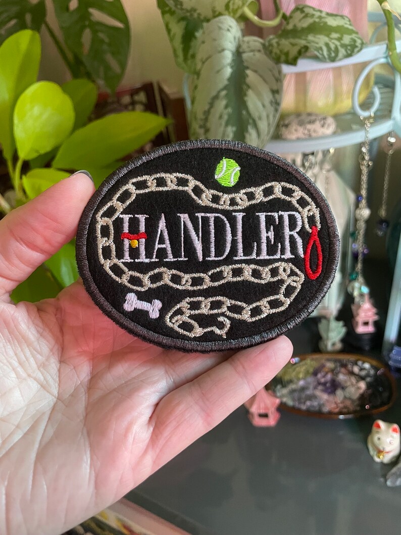 K&ouml;nnte beinhalten: Ein schwarzer gestickter Patch mit einem wei&szlig;en Kettenmuster und dem Wort "HANDLER" in wei&szlig;er Schrift. Ein rotes Halsband, ein wei&szlig;er Knochen und ein gr&uuml;ner Tennisball sind ebenfalls auf dem Patch gestickt.