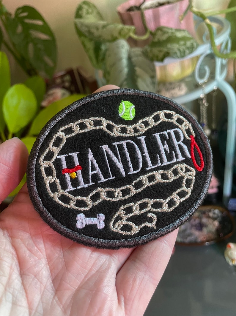 K&ouml;nnte beinhalten: Schwarzer gestickter Aufn&auml;her mit dem Wort "HANDLER" in Wei&szlig;, umgeben von einem Kettenmuster. Ein rotes Halsband, ein Knochen und ein gr&uuml;ner Tennisball sind ebenfalls auf den Aufn&auml;her gestickt.