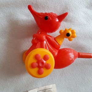 May include: Red and yellow plastic toy of a fox pulling a cart with a yellow wheel. The toy has a black eye and a small black dot on the nose. The toy has a label that reads "MOCKOCBCKAЯ ФАБРИКА ИГРУШЕК" in Cyrillic.
