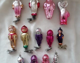 May include: A collection of 14 vintage glass ornaments shaped like people wearing space suits. The ornaments are in various colors, including pink, silver, gold, purple, and red. Each ornament has a metal clip on the bottom.