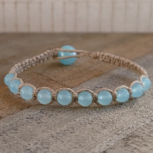 May include: A light blue gemstone bracelet with a knotted hemp cord. The bracelet has a single larger gemstone bead in the centre.