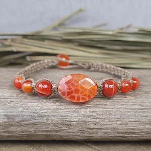May include: A bracelet made with natural hemp cord and orange carnelian beads. The bracelet features a large, oval-shaped carnelian bead in the center.