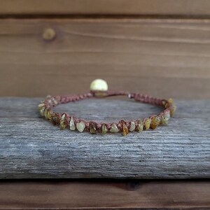 May include: A handmade bracelet featuring small, irregularly shaped amber stones strung on a brown cord. The bracelet is secured with a sliding knot and a small, round, light-colored bead. The bracelet is displayed on a weathered wooden surface.