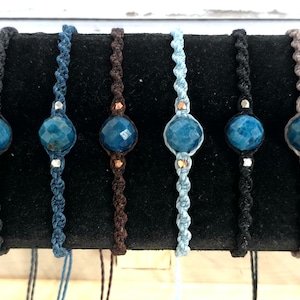 May include: A collection of seven macrame bracelets. Each bracelet features a faceted, blue gemstone bead. The macrame cords vary in color, including black, blue, brown, and gray. The beads are accented with small metal beads.