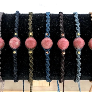 May include: A collection of seven macrame bracelets. Each bracelet features a large, pink gemstone bead. The macrame cords vary in color, including gray, navy, olive green, and brown. Small gold-colored beads accent the gemstone.