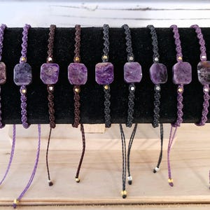 May include: Ten purple and white gemstone bracelets with adjustable cords. Each bracelet features a square-shaped gemstone with a unique pattern. The bracelets are displayed on a black velvet surface.