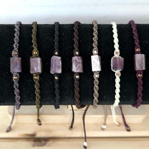 May include: Assortment of handmade macrame bracelets featuring rectangular amethyst stones. The bracelets are in various colors, including gray, olive green, black, brown, off-white, and purple. Each bracelet has a sliding knot closure.