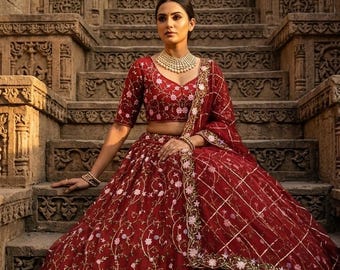 May include: A deep red lehenga set featuring a cropped blouse and a voluminous skirt, both adorned with intricate white and light purple floral embroidery. The ensemble includes a matching sheer dupatta with a gold grid pattern and floral borders.